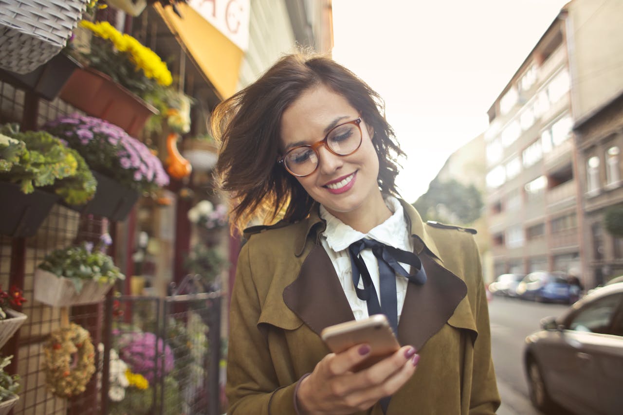 A smiling woman uses her phone outside a colorful urban flower shop on a sunny day.